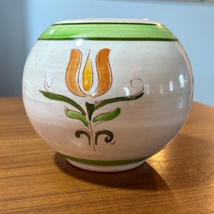 May include: A round, off-white ceramic vase with a hand-painted floral design. The vase features a green band at the top and bottom, with an orange and yellow tulip-like flower in the center. The vase is sitting on a wooden surface.
