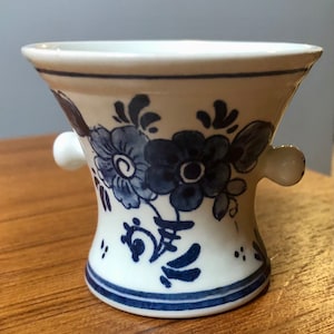 May include: A small, white ceramic vase with a flared top and two small handles. The vase is decorated with blue floral patterns and accents. The base and rim have blue stripes. The vase is sitting on a wooden surface.