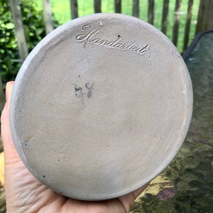 May include: The base of a ceramic pot with the word "Handarbeit" inscribed on it. The pot is light grey and has a smooth finish.
