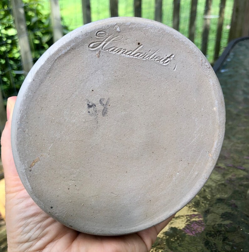 May include: The base of a ceramic pot with the word "Handarbeit" inscribed on it. The pot is light grey and has a smooth finish.