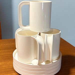 May include: A set of four off-white mugs with large, rectangular handles, stacked on a matching circular tray. The mugs are cylindrical with a smooth, glossy finish. The tray has a series of concentric rings. The background is a light blue wall and a wooden table.