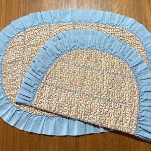 May include: Two-piece set of floral print mats with a light blue ruffled border. The mats have a cream background with a pattern of small red, blue, and green flowers. The border is a solid light blue with white polka dots.