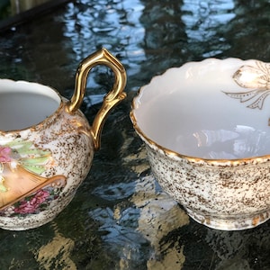 May include: A vintage cream pitcher and sugar bowl set. The white porcelain pieces have a speckled gold and brown pattern. The pitcher has a gold handle and floral accents. The sugar bowl has a scalloped edge and gold bow design.