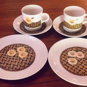 May include: A set of white ceramic teacups and saucers. The cups and saucers feature a woven basket design with a daisy flower in the center. The set includes two cups and four saucers.