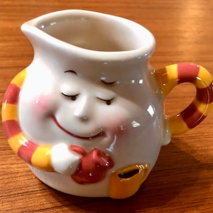 May include: A whimsical, off-white ceramic creamer with a smiling face design. The creamer features a red, yellow, and orange striped handle and a matching design around the neck. A small red teapot is held by the creamer's face, and a yellow coffee cup is on the side.