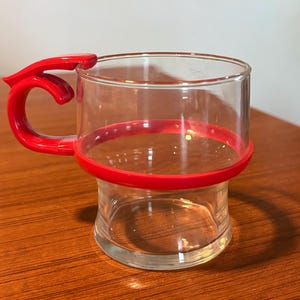 May include: A clear glass mug with a red handle and a red band around the middle. The mug sits on a wooden surface. The handle has a unique, curved design.