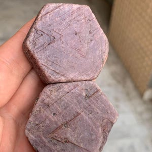 May include: Two hexagonal, raw, opaque, dusty-pink gemstones with visible surface striations and natural inclusions. The stones have a flat, matte texture and irregular edges, displaying a distinct six-sided geometric shape with earthy tones.