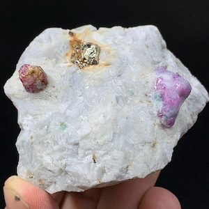 May include: A white rock formation with three embedded gemstones. Two are a deep pink-red color, and one is a gold-colored, metallic cube. The rock has a rough, textured surface and is held in a hand.