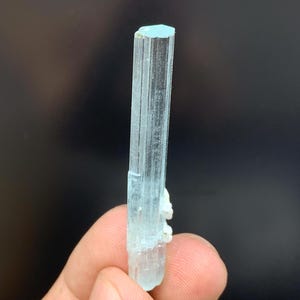 May include: A pale blue aquamarine crystal with a hexagonal top. The crystal is held between fingers, showcasing its translucent quality and natural, elongated form. The crystal has a light blue hue and a slightly rough texture.
