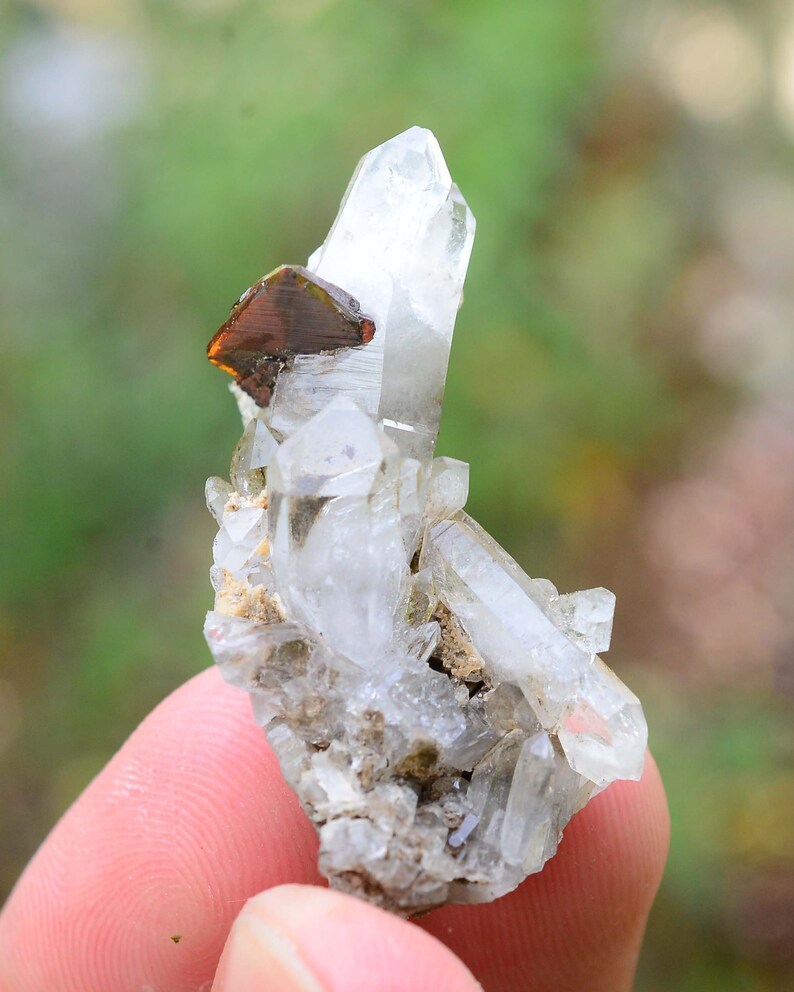 May include: A cluster of clear quartz crystals with a single, large, reddish-brown crystal on top. The crystals are on a bed of grey rock.