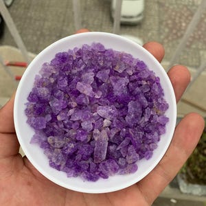 May include: A white ceramic bowl filled with raw, uncut amethyst crystals. The crystals are a deep purple color with some lighter, translucent areas. The bowl is held in a hand, with a blurred outdoor background.