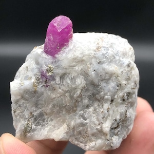 Könnte beinhalten: Ein leuchtend rosa Rubin-Kristall, eingebettet in eine raue, weiße und graue Gesteinsmatrix. Der Rubin hat eine tiefe, satte rosafarbene Farbe und eine scharfe, facettierte Form. Die Gesteinsmatrix ist mit kleinen, weißen Kristallen bedeckt und hat eine raue, strukturierte Oberfläche.