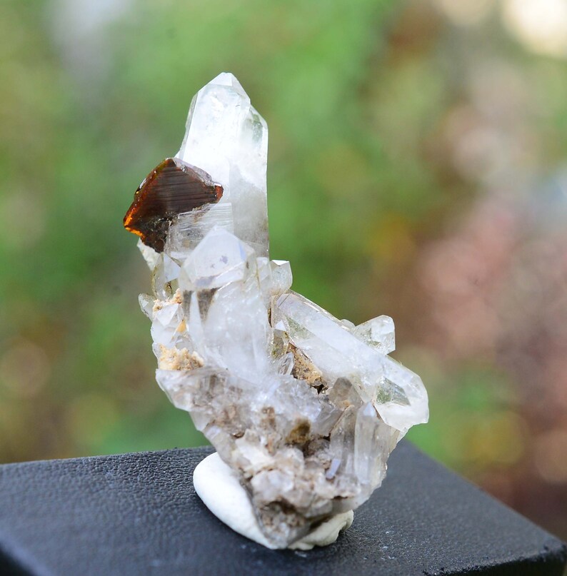 May include: A cluster of clear quartz crystals with a brown, metallic mineral inclusion. The crystals are arranged in a cluster on a white base.