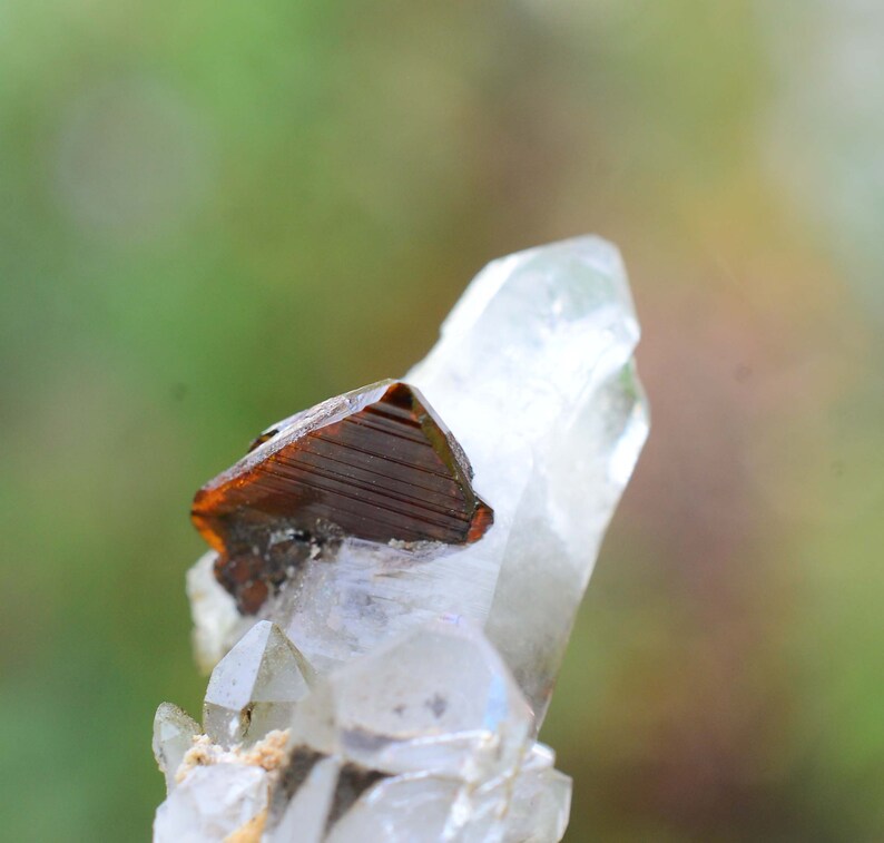 May include: A clear quartz crystal with a brown, faceted, mineral inclusion. The crystal is pointed at the top and has a rough, natural texture.