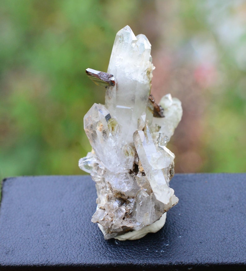 May include: A cluster of clear quartz crystals with a small piece of brown haematite. The crystals are arranged in a vertical formation, with the largest crystal at the top. The crystals are set on a dark background.