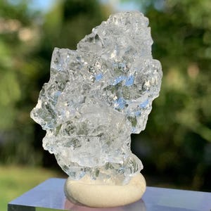Può includere: Una scultura cristallina trasparente con una superficie ruvida e strutturata. La scultura è montata su una piccola pietra arrotondata bianco sporco, che è posta su una base riflettente color argento. Lo sfondo è sfocato, con fogliame verde e cielo blu.