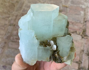 May include: A cluster of light blue aquamarine crystals with a rough, natural texture. The crystals are clustered together in a random formation, with some crystals larger than others. The crystals are translucent and have a slight green tint.