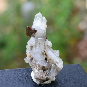 May include: A cluster of clear quartz crystals with a reddish-brown mineral inclusion. The crystals are arranged in a pointed formation, with the largest crystal at the top. The crystals are set on a grey, rocky base.
