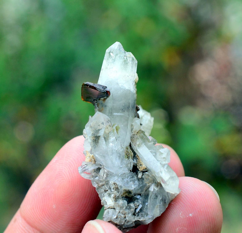 May include: A small cluster of clear quartz crystals with a brown mineral inclusion. The crystals are clustered together on a grey matrix.