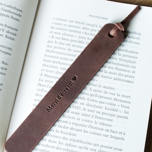 Peut inclure: Un marque-page en cuir marron avec le texte "Merci d'être toi" et un symbole de cœur. Le marque-page est rectangulaire avec une boucle en haut et est placé sur un livre ouvert.