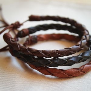 May include: Three braided leather bracelets in shades of brown and black. The bracelets are made of woven leather strips, with a rustic and textured appearance. The bracelets are likely adjustable, with a leather tie closure.