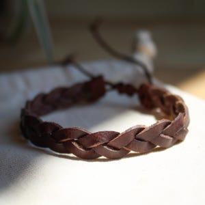 Könnte beinhalten: Ein geflochtenes Armband aus braunem Leder. Das Armband besteht aus mehreren Strängen Leder, die miteinander verflochten sind. Das Armband hat eine dunkelbraune Farbe und scheint handgefertigt zu sein. Es liegt auf einer weißen Oberfläche.