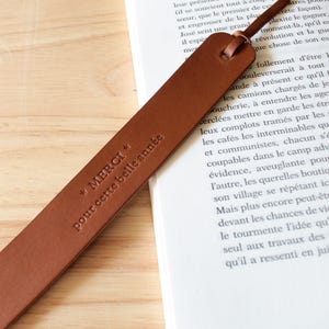 Peut inclure: Un marque-page en cuir marron avec le texte en relief "MERCI pour cette belle année" et une boucle en cuir. Le marque-page est posé sur un livre avec du texte imprimé.