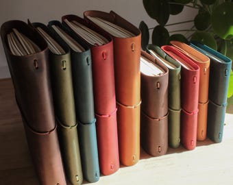 Peut inclure: Une collection de dix carnets reliés en cuir disposés en rangée, présentant diverses couleurs dont le brun foncé, le vert olive, le bleu sarcelle, le rouge, le beige et le bleu. Chaque carnet est maintenu par un cordon élastique fin.