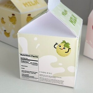 May include: A white cardboard milk carton with a green melon character on the front. The carton has a nutrition facts label on the side and the text "CNDLNYC" on the bottom.