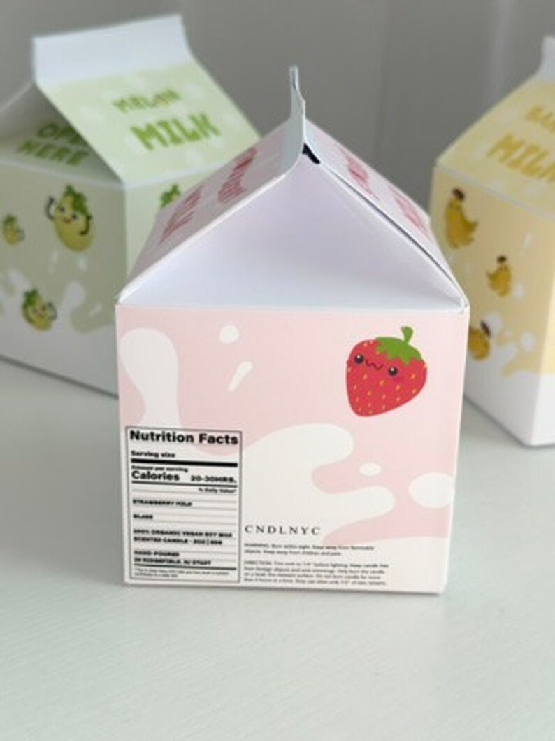 May include: A pink and white milk carton candle with a strawberry illustration and the text "Nutrition Facts" and "CANDLNYC".