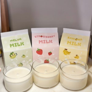 May include: Three glass candle jars with white wax. The jars are labeled "Melon Milk", "Strawberry Milk", and "Banana Milk". Each label features a cartoon fruit with a smiling face.