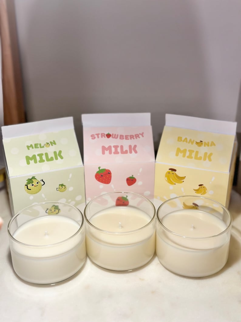 May include: Three glass candle jars with white wax. The jars are labeled "Melon Milk", "Strawberry Milk", and "Banana Milk". Each label features a cartoon fruit with a smiling face.