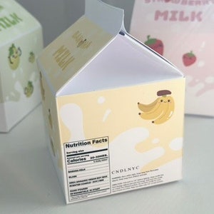 May include: A yellow cardboard box shaped like a milk carton with a nutrition facts label and the text "CNDLNYC". The box has a cartoon banana with a smiling face on the front.