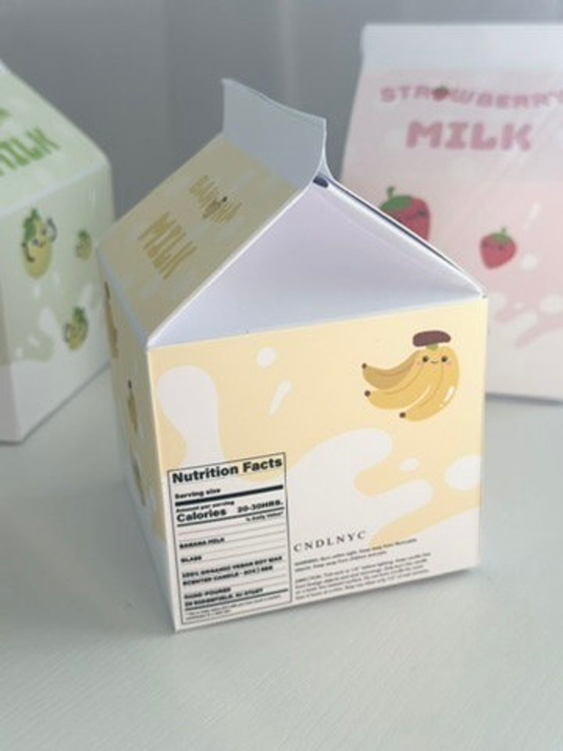May include: A yellow cardboard box shaped like a milk carton with a nutrition facts label and the text "CNDLNYC". The box has a cartoon banana with a smiling face on the front.