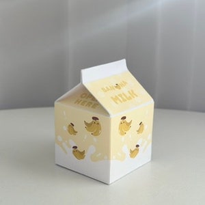 May include: A small cardboard box shaped like a milk carton with a yellow and white design. The box has a cartoon image of bananas and the words "Banana Milk" on the front. The box says "Open Here" on the side.