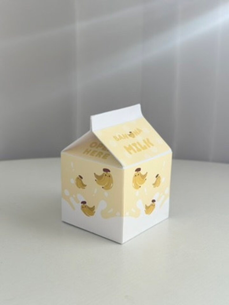 May include: A small cardboard box shaped like a milk carton with a yellow and white design. The box has a cartoon image of bananas and the words "Banana Milk" on the front. The box says "Open Here" on the side.