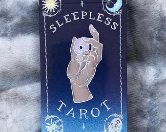 May include: A blue box with a white border and the text "Sleepless Tarot" in white. The box features a hand holding a purple eye with a yellow iris. The box has a white border with a sun, moon, and other symbols.