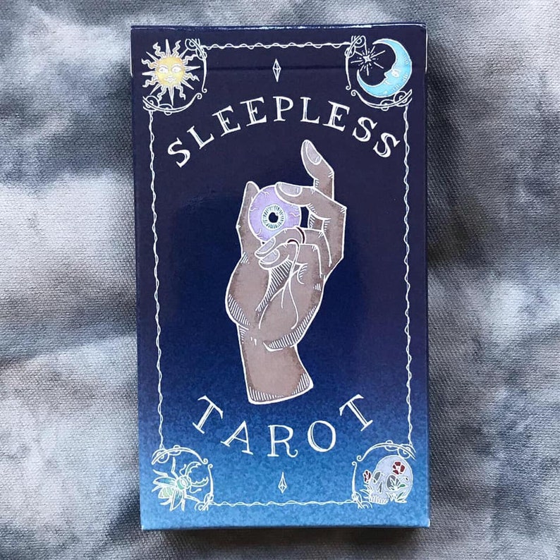 May include: A blue box with a white border and the text "Sleepless Tarot" in white. The box features a hand holding a purple eye with a yellow iris. The box has a white border with a sun, moon, and other symbols.