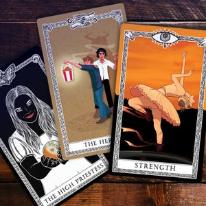 May include: Three tarot cards with black borders and ornate designs. The first card is black with a white illustration of a woman holding a sphere. The text "THE HIGH PRIESTESS" is at the bottom. The second card is brown with a cartoon illustration of two men. The text "THE HERMIT" is at the bottom. The third card is orange with a black and white illustration of a woman in a white tutu. The text "STRENGTH" is at the bottom.