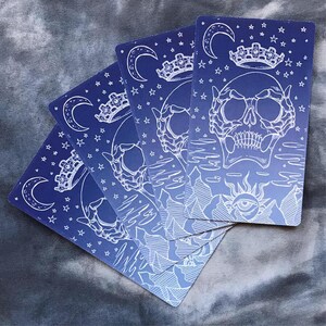 May include: Five tarot cards with a blue and white gradient background. Each card features a white skull with a crown, a crescent moon, and stars. The cards are arranged in a fan shape.