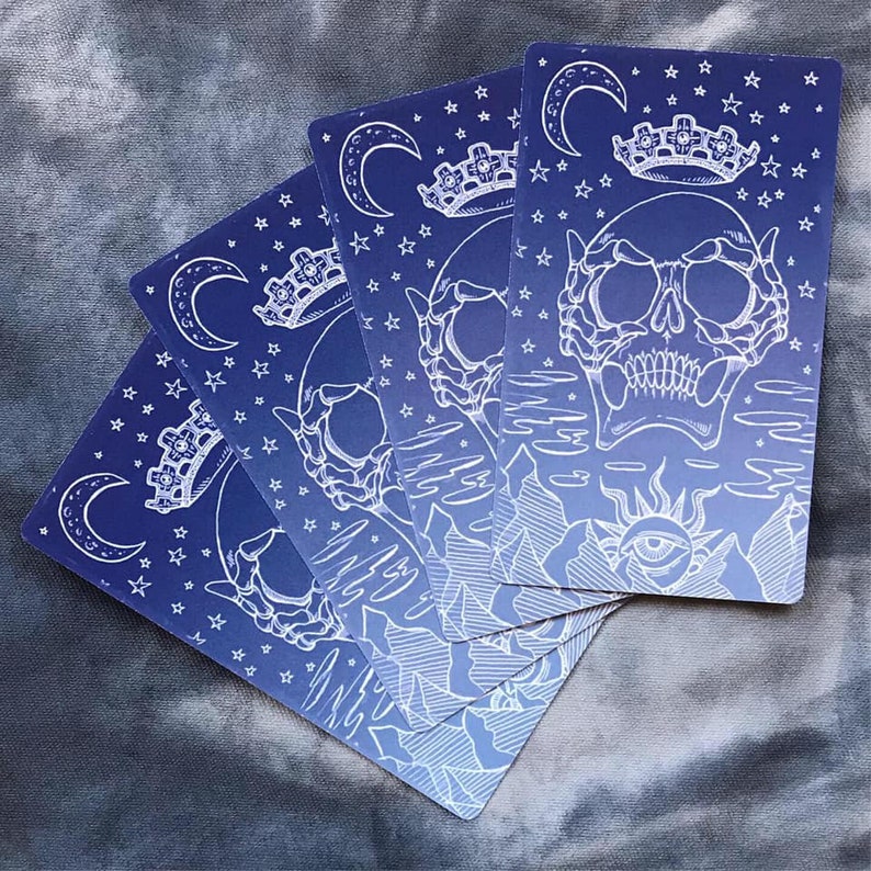May include: Five tarot cards with a blue and white gradient background. Each card features a white skull with a crown, a crescent moon, and stars. The cards are arranged in a fan shape.