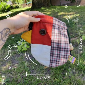 Op de afbeelding: Een stoffen buidel met trekkoord in de vorm van een vogel, met een rood bovendeel, een wit onderdeel, een gele snavel en een bruin geruit zijpaneel. Een donkere knoop dient als oog. De buidel meet ongeveer 17 cm bij 22 cm.
