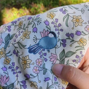 Può includere: Un tessuto di colore chiaro con un motivo floreale ripetuto di narcisi gialli, fiori viola a forma di campana e fiori rosa, con un'illustrazione centrale di un piccolo uccello blu con testa nera e una macchia bianca sulla guancia.