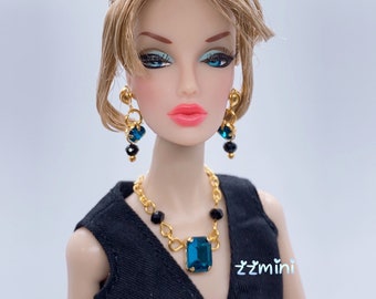 May include: A doll wearing a black dress with a gold chain necklace and a large teal gemstone pendant. The doll has blonde hair styled in a bouffant and is wearing gold earrings with teal and black gemstones.