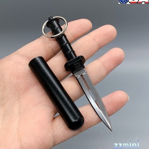 Può includere: Un coltellino nero per portachiavi con una lama argentata e un fodero nero. Il coltellino ha un anello nella parte superiore per essere attaccato a un portachiavi.