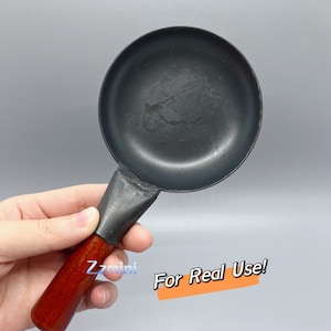 May include: A small, black, cast iron frying pan with a wooden handle. The pan is about 6 inches in diameter and has a slightly rounded bottom. The handle is about 5 inches long and has a smooth, brown finish. The pan is labeled "For Real Use!" in orange text.