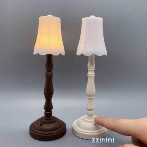 Pu&ograve; includere: Due lampade da tavolo in miniatura con paralumi bianchi. La lampada a sinistra ha una base marrone e la lampada a destra ha una base bianca. La lampada a destra ha un interruttore sulla base.