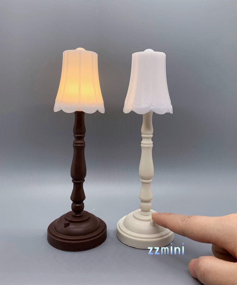 Pu&ograve; includere: Due lampade da tavolo in miniatura con paralumi bianchi. La lampada a sinistra ha una base marrone e la lampada a destra ha una base bianca. La lampada a destra ha un interruttore sulla base.