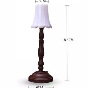 Pu&ograve; includere: Una lampada da tavolo in legno marrone con un paralume in tessuto bianco. La lampada &egrave; alta 18,5 cm, larga 3 cm e ha un diametro di 6 cm alla base.
