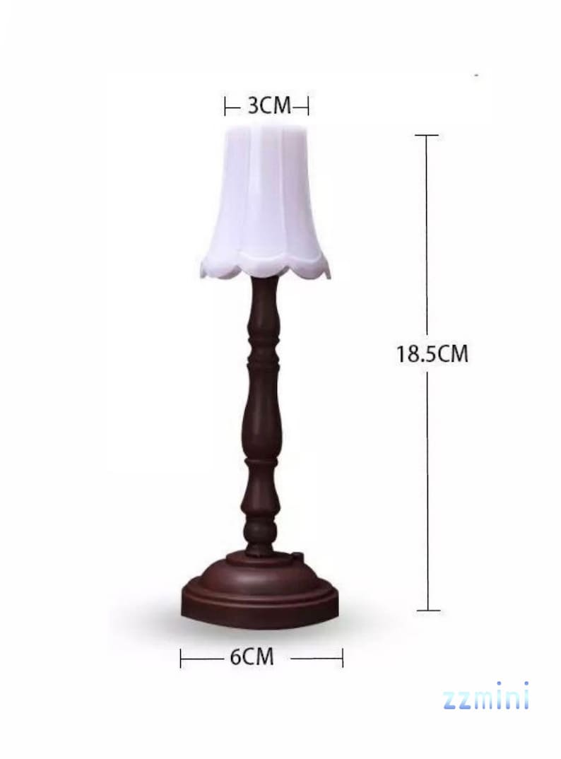 Pu&ograve; includere: Una lampada da tavolo in legno marrone con un paralume in tessuto bianco. La lampada &egrave; alta 18,5 cm, larga 3 cm e ha un diametro di 6 cm alla base.